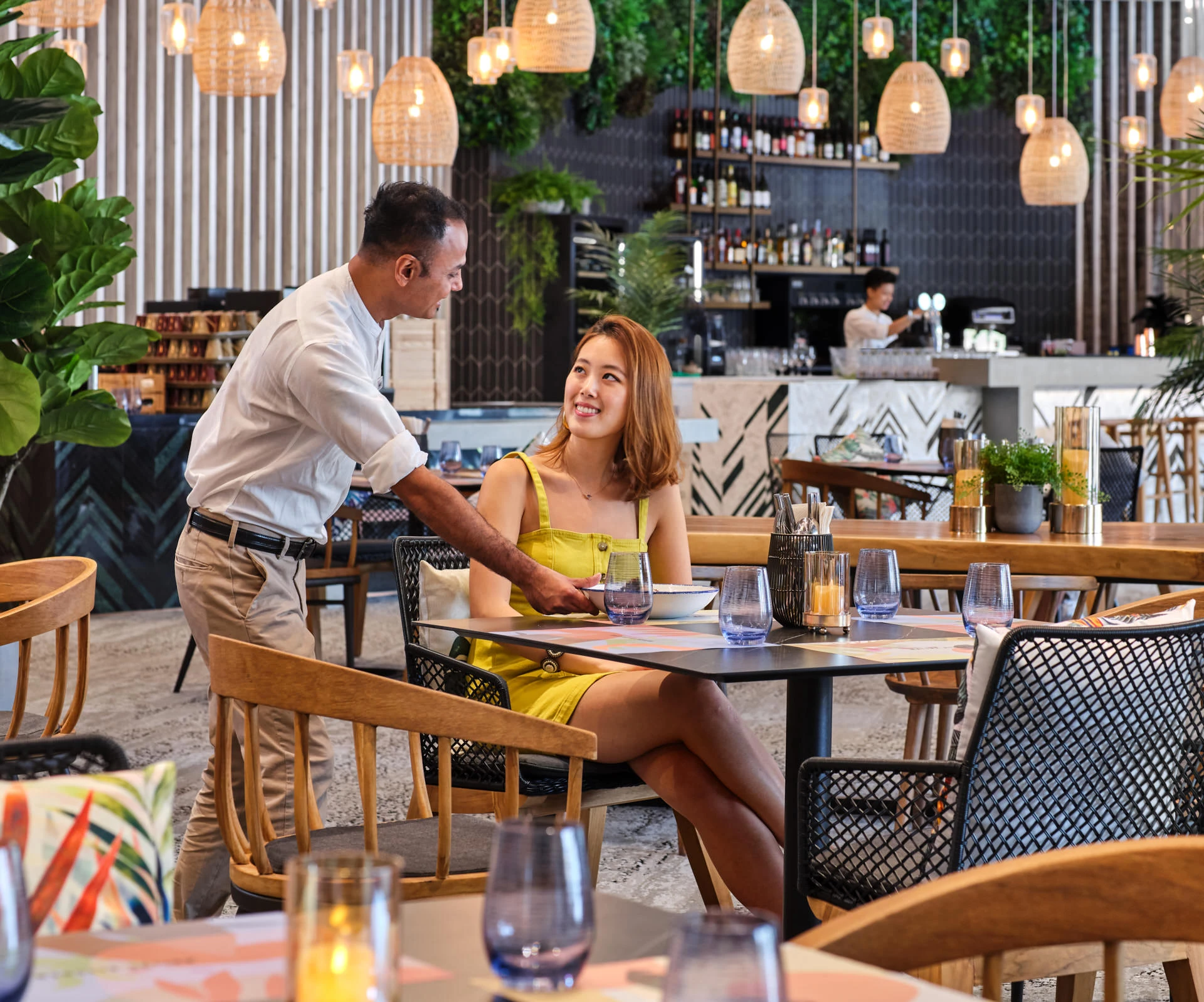 Staff serving guest at restaurant table with bar and rattan pendants behind — Village Hotel Sentosa lifestyle photography