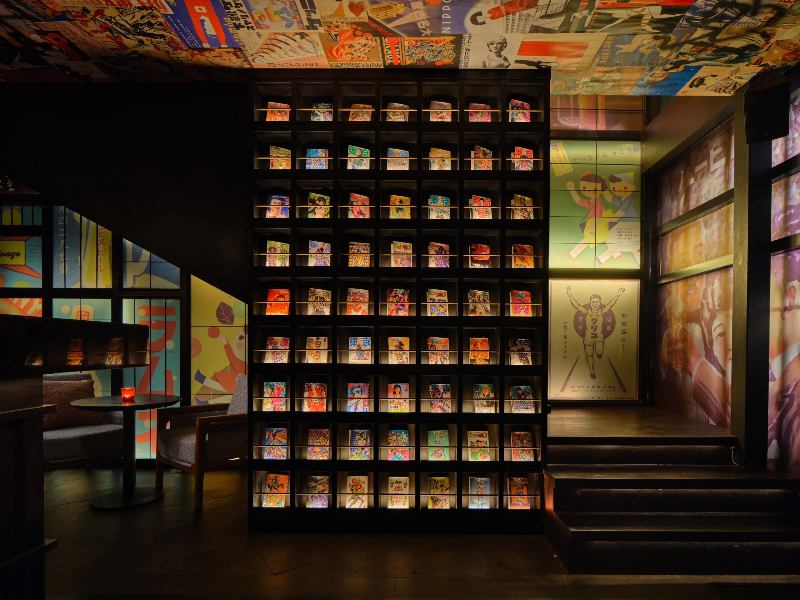 Dark entrance with a tall backlit bookshelf displaying colorful Japanese manga and books, collage-covered ceiling, and illuminated vintage poster panels.