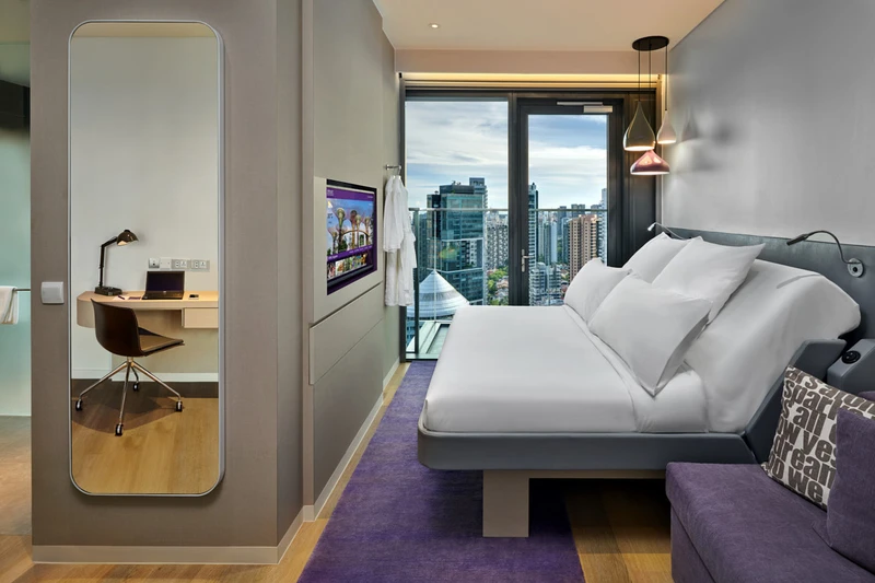 Modern hotel room with a platform bed, white duvet, purple carpet and sofa, full-length mirror, wall-mounted TV, desk area, and high-rise city views.