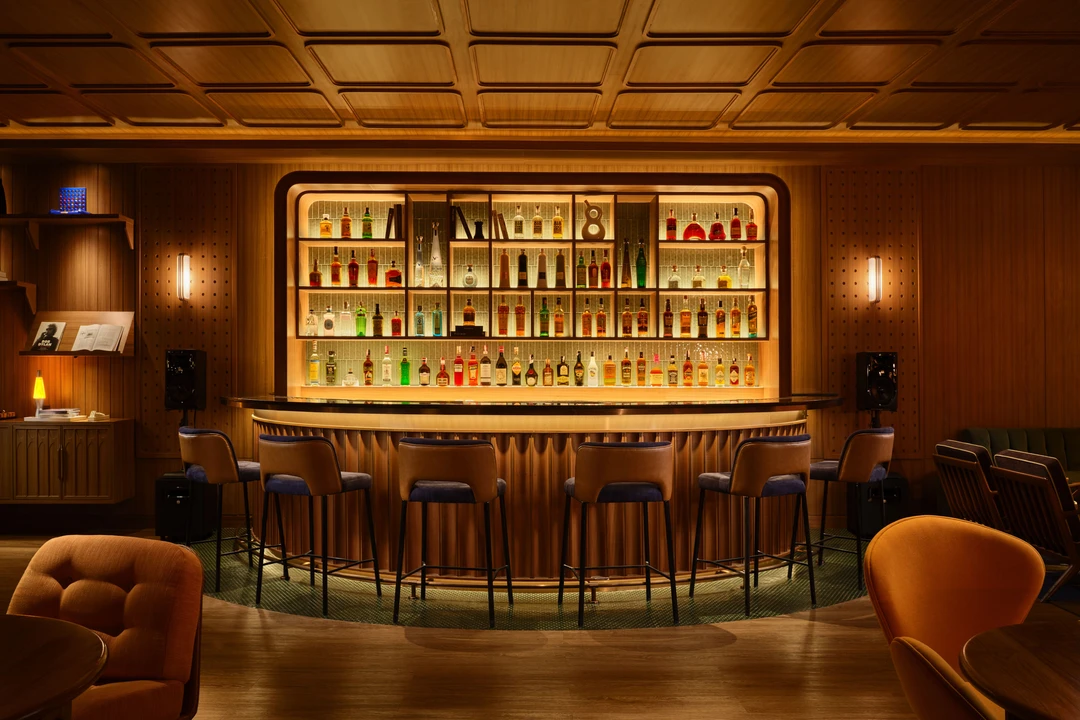 Curved wood-paneled bar with backlit bottle display, leather stools, and coffered ceiling in warm ambient lighting