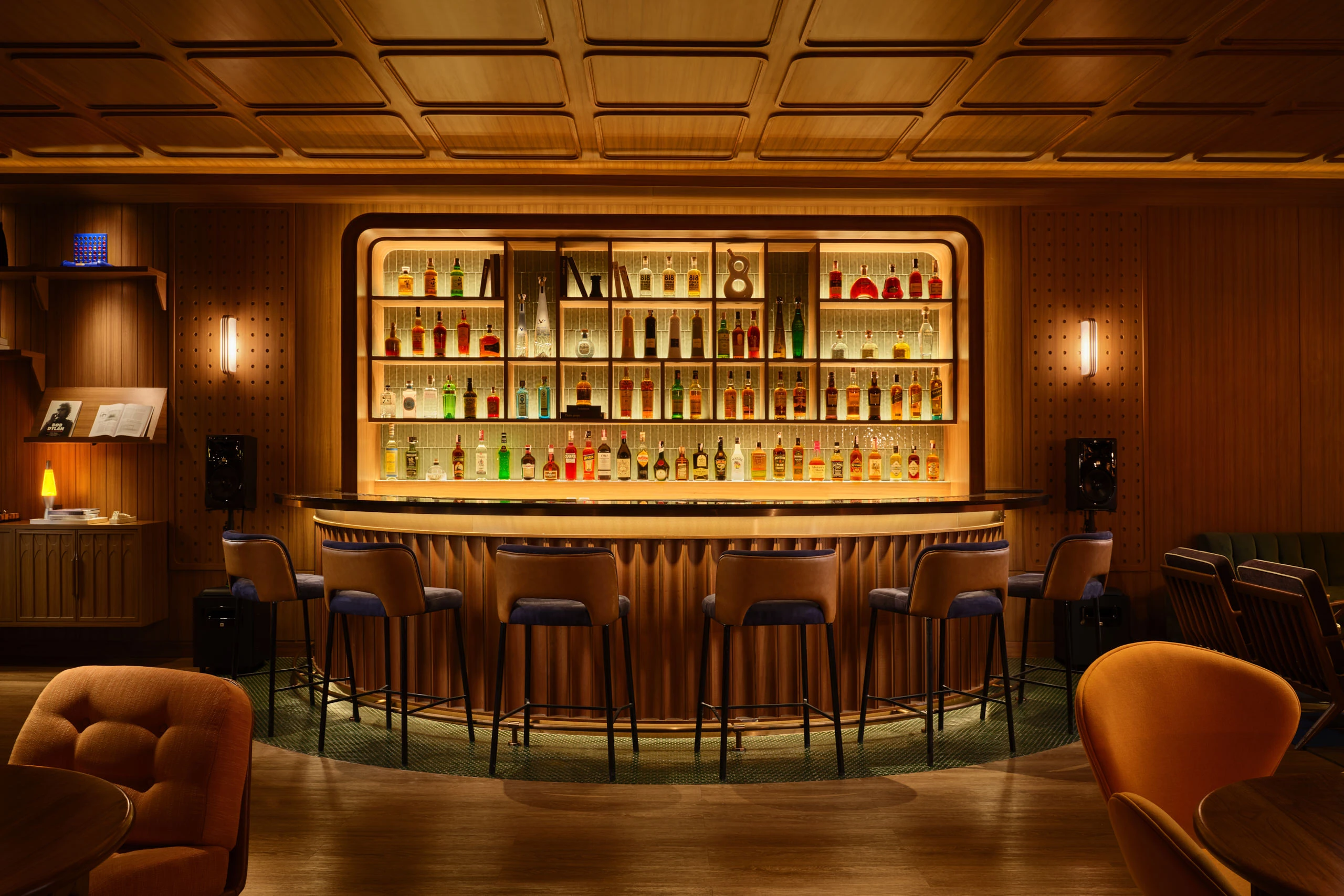 Curved wood-paneled bar with backlit bottle display, leather stools, and coffered ceiling in warm ambient lighting