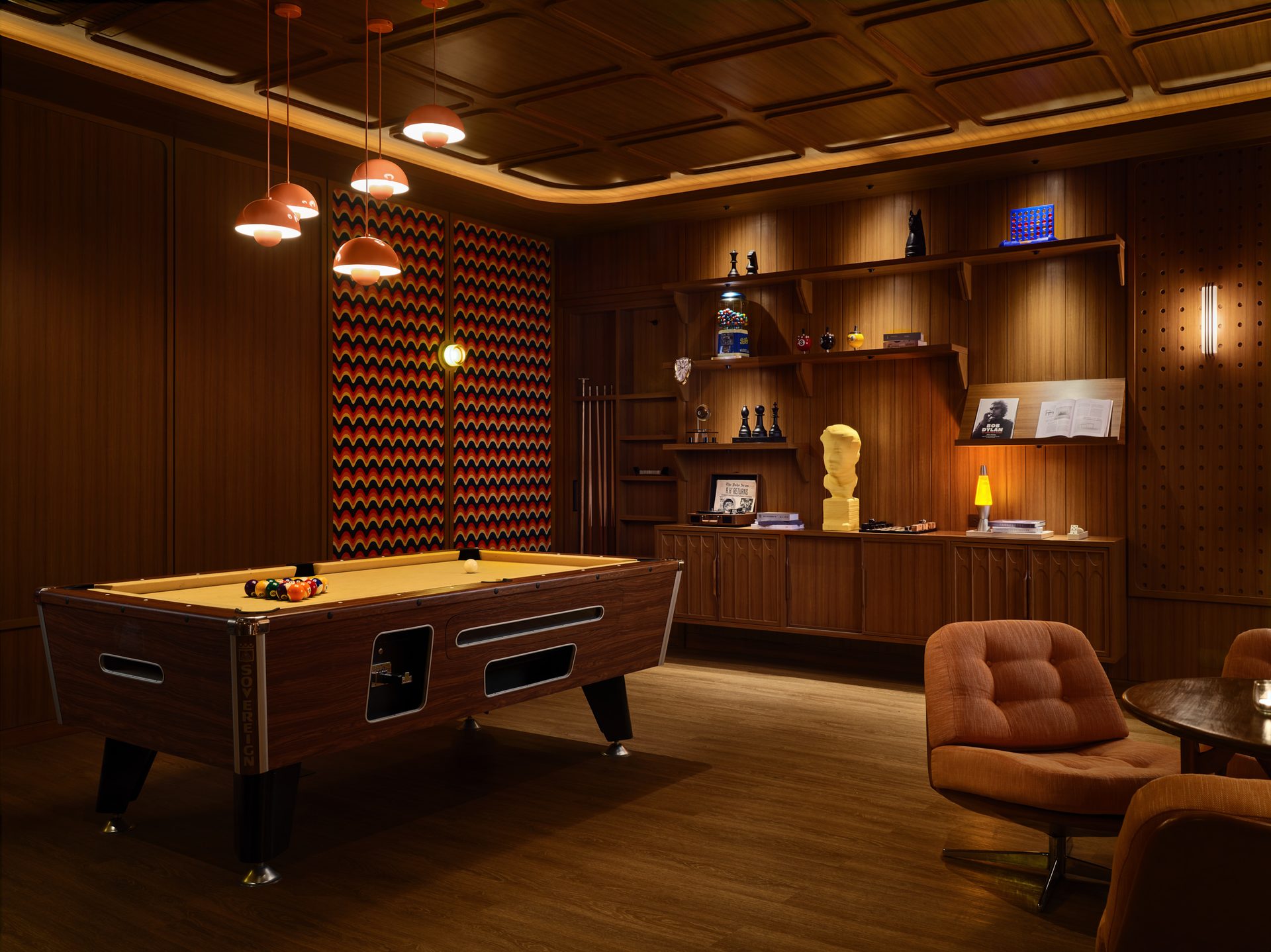 Pool table in wood-paneled game room with chevron accent wall, copper pendant lights, and vintage furnishings