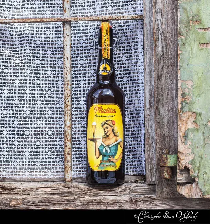 Mielita birra artigianale bottle at a rustic weathered window with lace curtain
