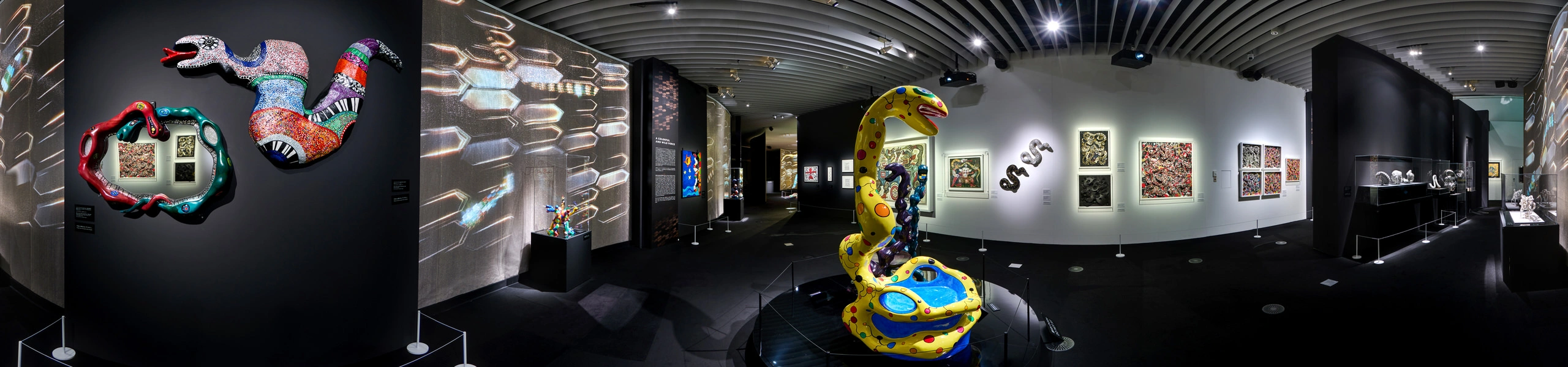 Panoramic gallery with colourful mosaic snake wall sculptures, large spotted serpent floor sculpture, and framed artworks on white walls