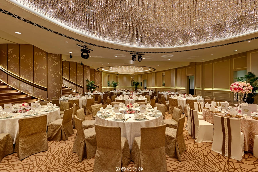 Grand ballroom with a massive crystal chandelier ceiling, round banquet tables dressed in white linen and gold chair covers, and a curved staircase at left.