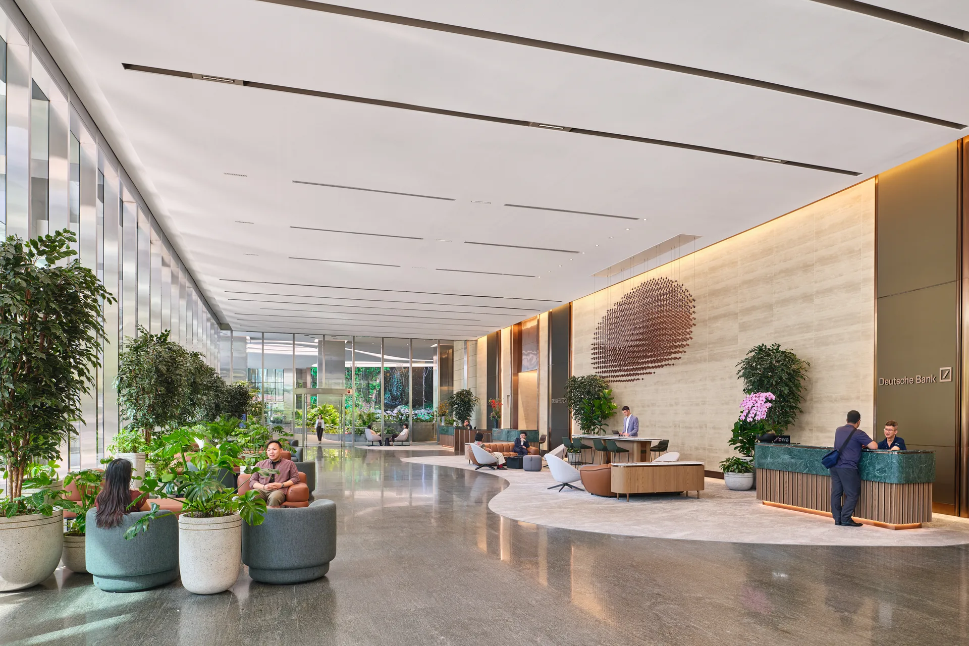 Grand commercial tower lobby with high ceilings, polished floors, large potted plants, circular metal wall sculpture, seating areas, and a reception desk.