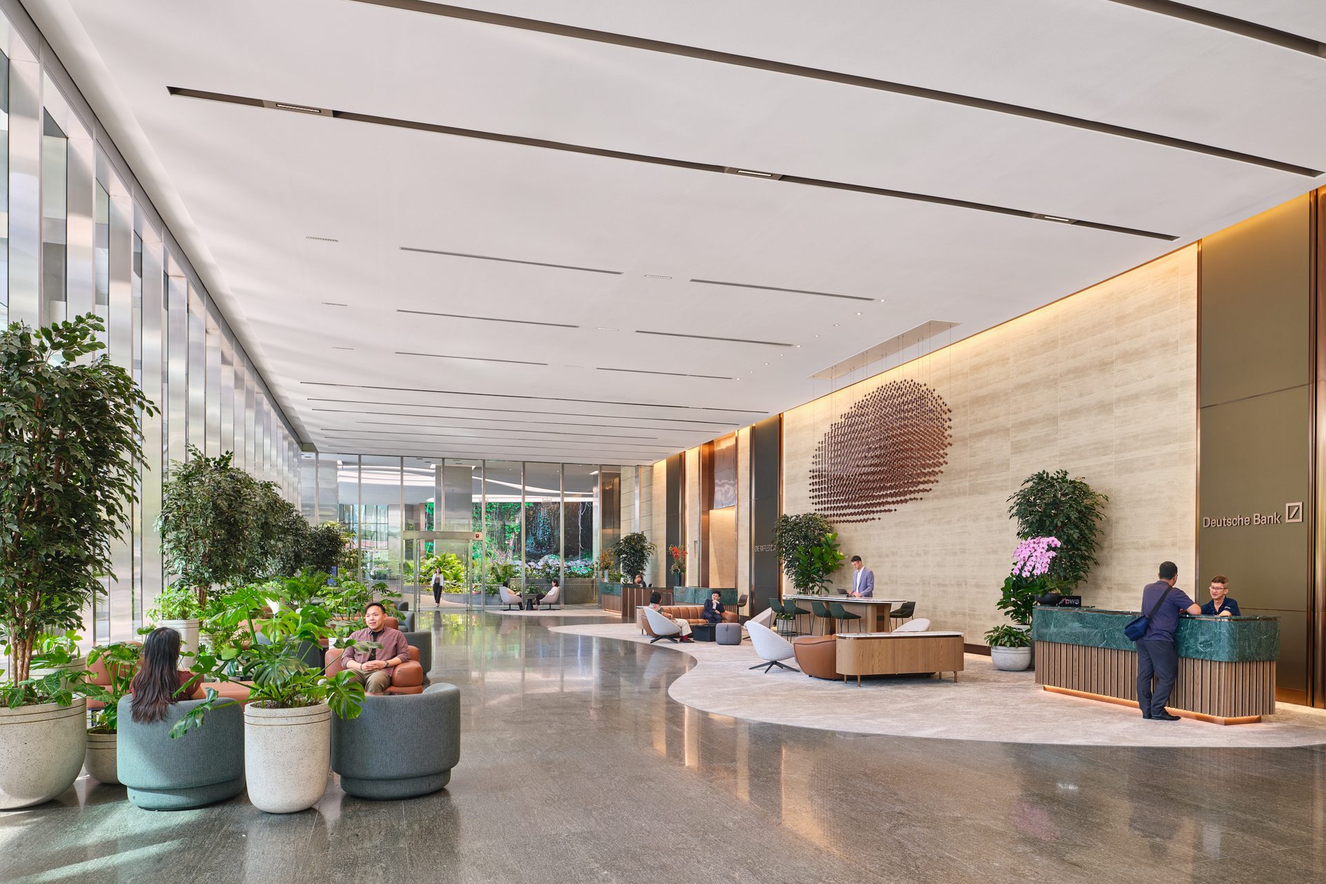 Grand commercial tower lobby with high ceilings, polished floors, large potted plants, circular metal wall sculpture, seating areas, and a reception desk.