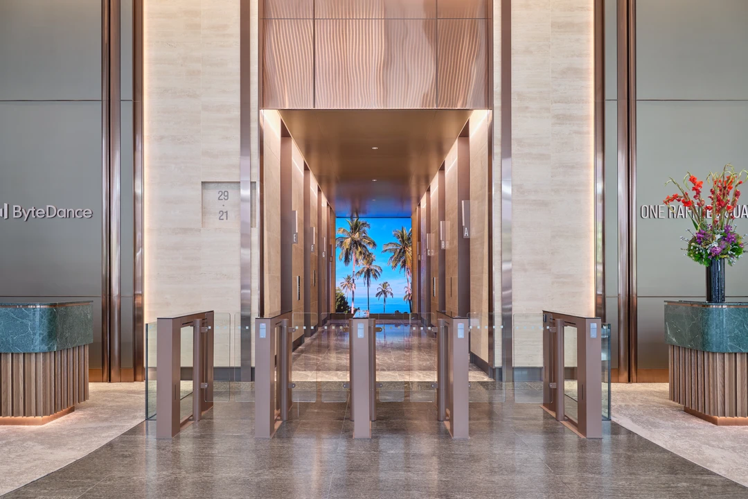 Modern office tower lobby with marble floors, bronze turnstile gates, palm tree mural visible through the entrance, and recessed ceiling lighting.
