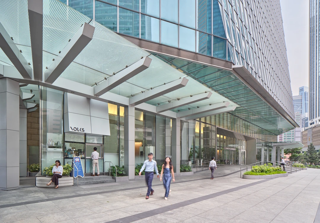 Glass-and-steel office tower entrance canopy with pedestrians walking on grey paving beneath angular structural beams and surrounding high-rise buildings.