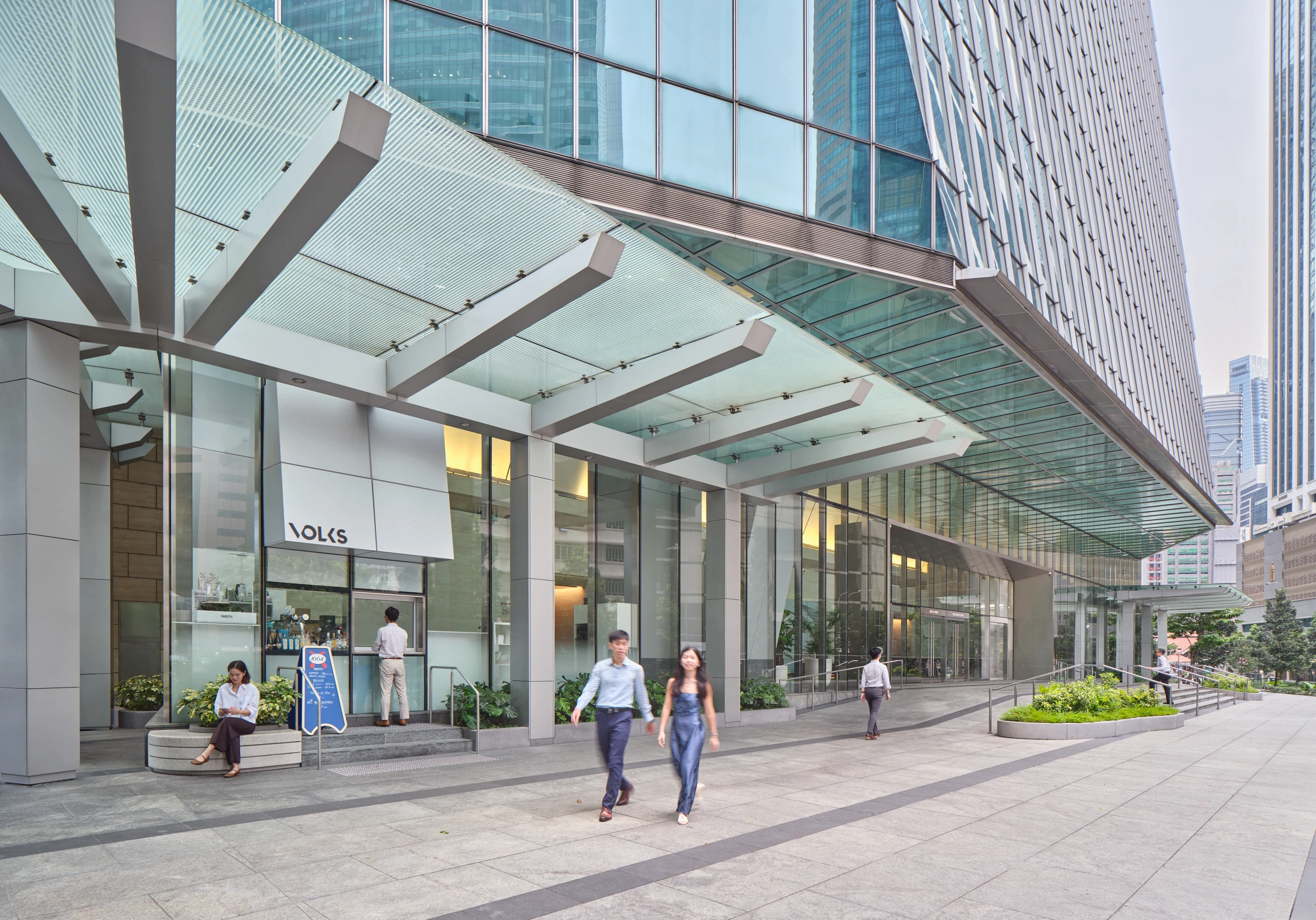 Glass-and-steel office tower entrance canopy with pedestrians walking on grey paving beneath angular structural beams and surrounding high-rise buildings.