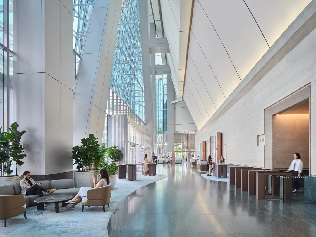 Double-height office lobby with soaring glass atrium ceiling, potted fiddle-leaf fig trees, grey lounge seating, polished floors, and turnstile gates at the reception area.