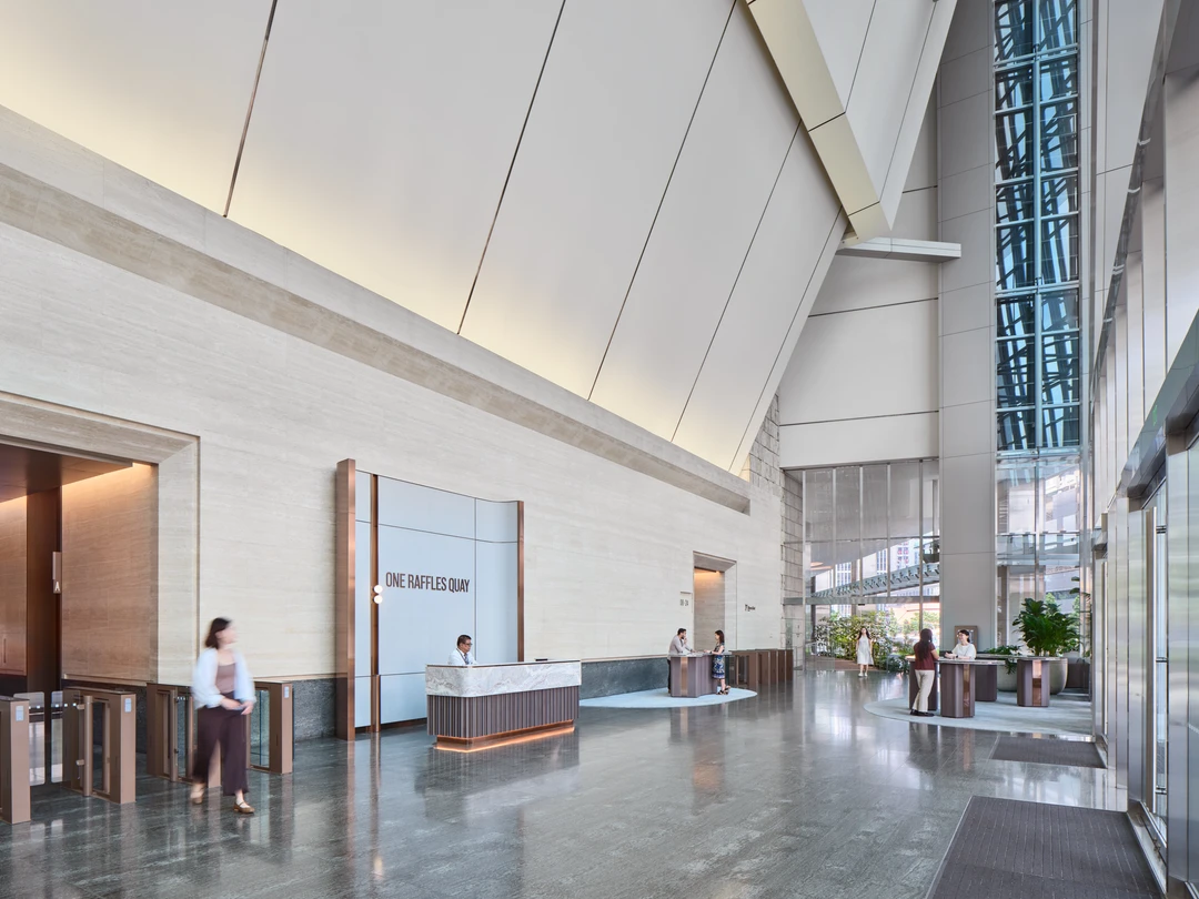 Grand lobby interior with angular white ceiling panels, stone-clad walls, a reception desk bearing the One Raffles Quay sign, and a glass atrium at right.