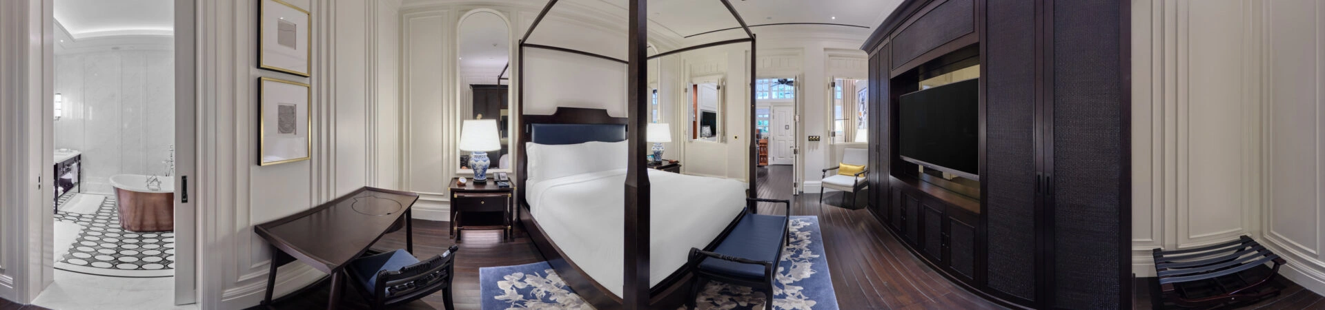 Panoramic view of a colonial-style hotel suite with dark four-poster bed, white linens, blue-and-white porcelain lamps, dark wood floors, and an adjoining marble bathroom.