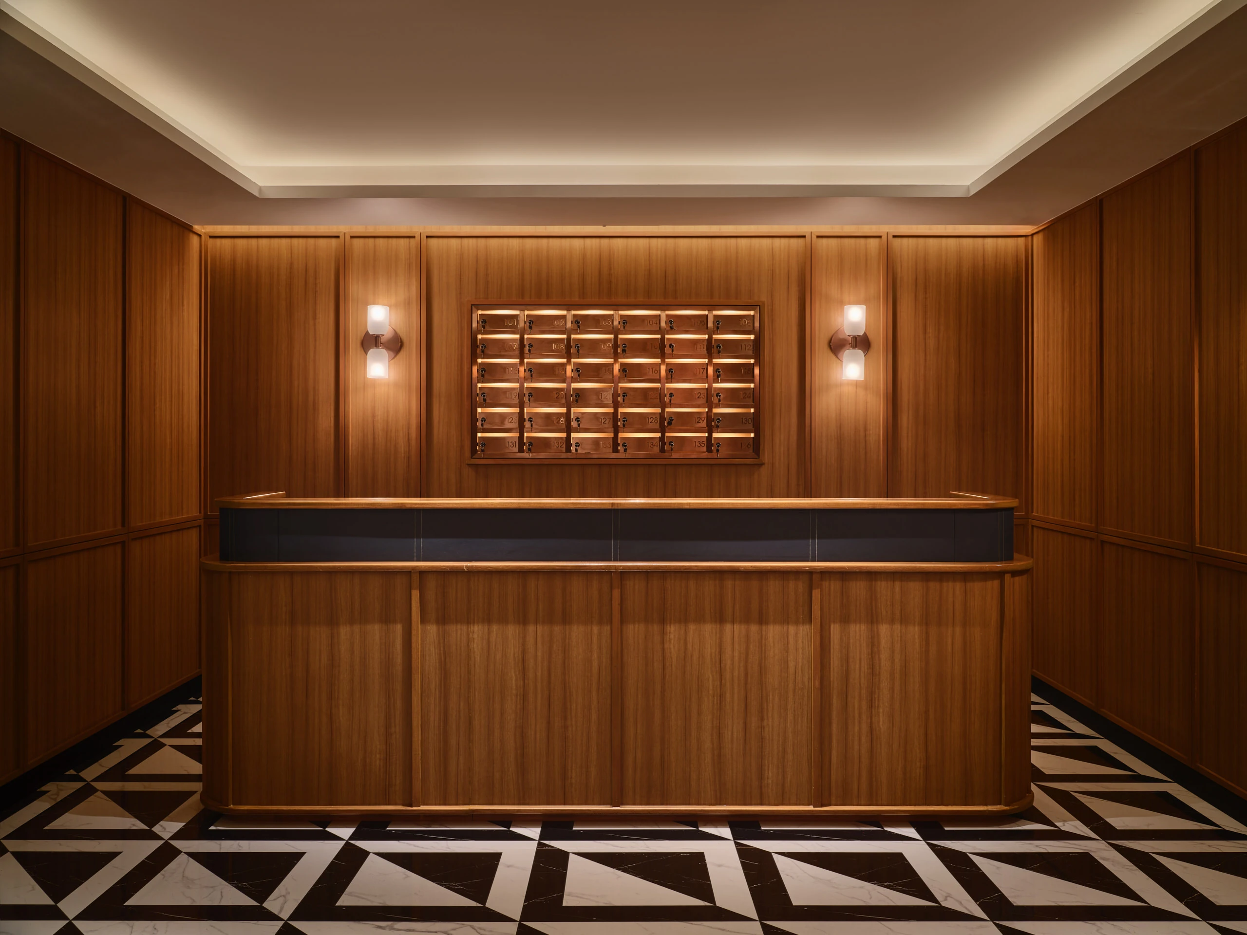 Reception desk with warm wood paneling, brass mailbox grid, geometric black-and-white floor tiles, and wall sconces