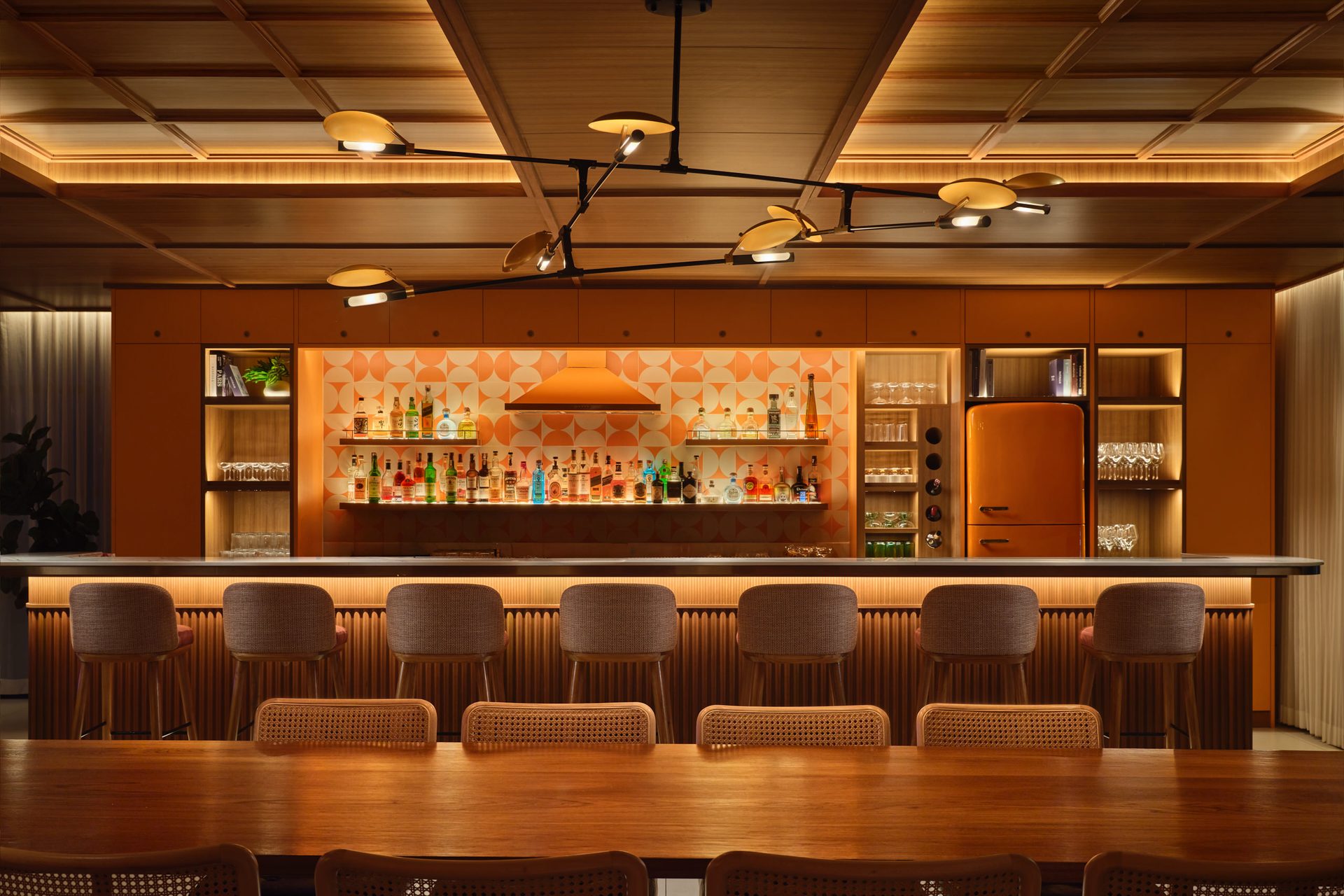 Long bar with cane-backed stools, orange geometric backsplash, retro fridge, and modern sputnik chandelier overhead