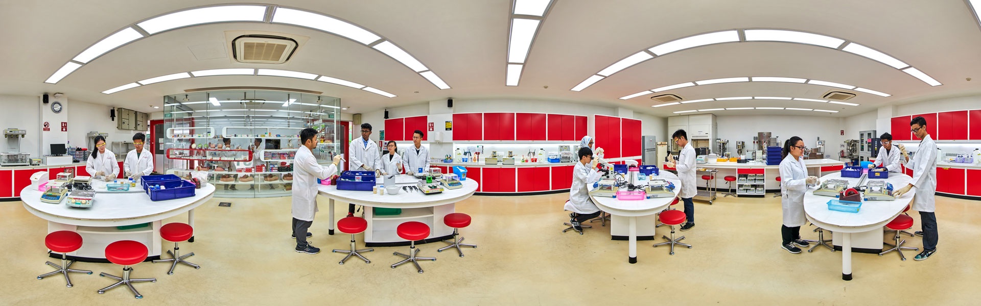 Panoramic view of a pharmacy compounding laboratory with students in white lab coats working at round white tables, red stools, and red cabinet walls.