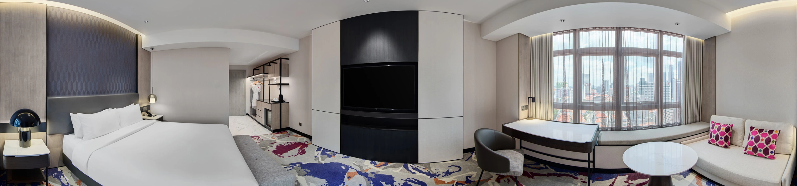 Panoramic view of a hotel room with white king bed, bold multicoloured carpet, wall-mounted TV, curved window sofa with pink cushions, and city views.