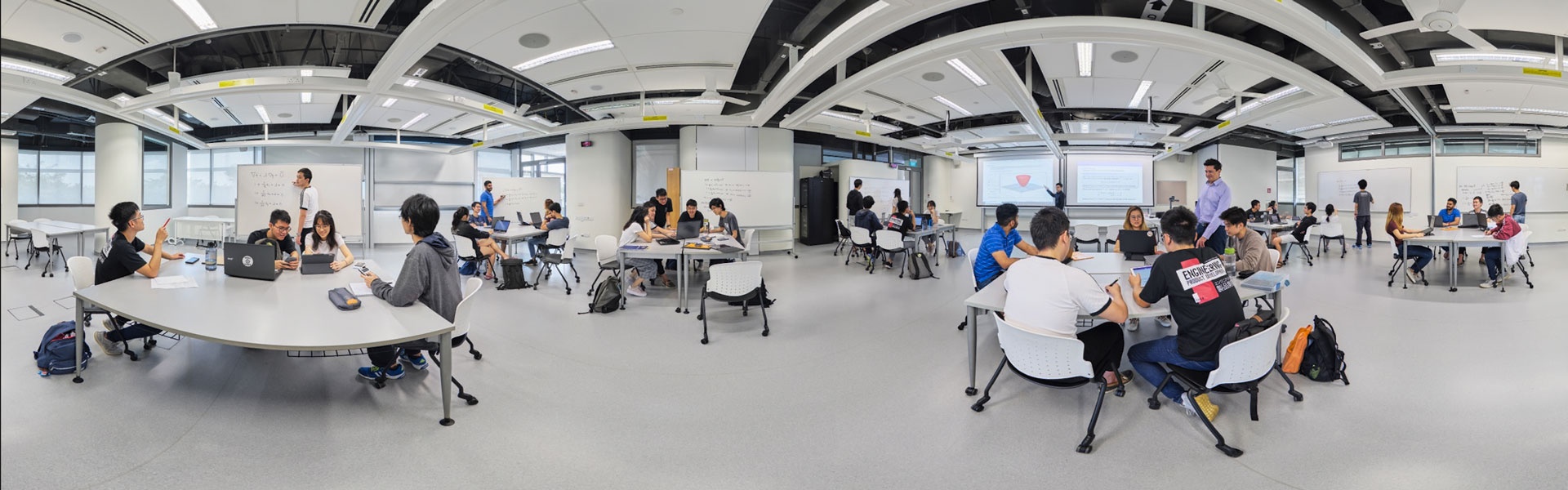 Panoramic view of a university cohort classroom with students working in groups at curved white tables, whiteboards, exposed ceiling ducts, and a projector screen.