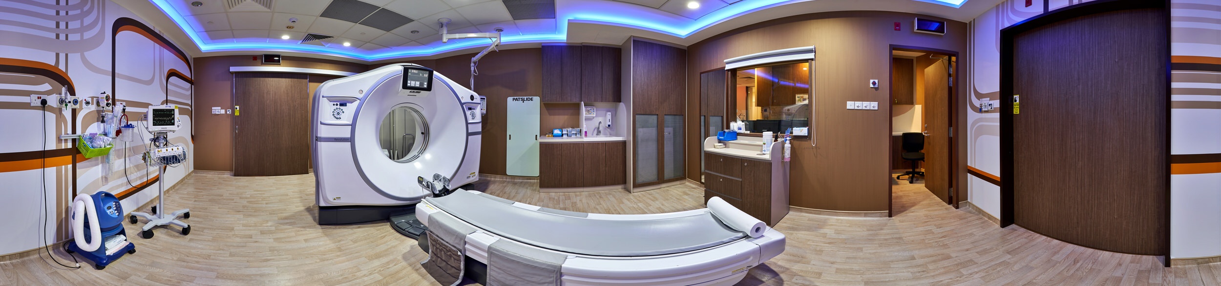 Panoramic view of a hospital CT scan room with white scanner, patient bed, medical monitoring equipment, dark wood paneling, and blue accent lighting.