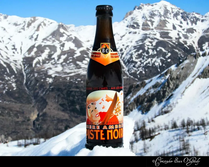 Piste Noire amber beer bottle nestled in snow with Alpine peaks behind