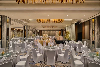 Wide view of a hotel grand ballroom with white banquet rounds, marble columns, patterned carpet, a lit stage backdrop, and gold pendant chandeliers overhead.