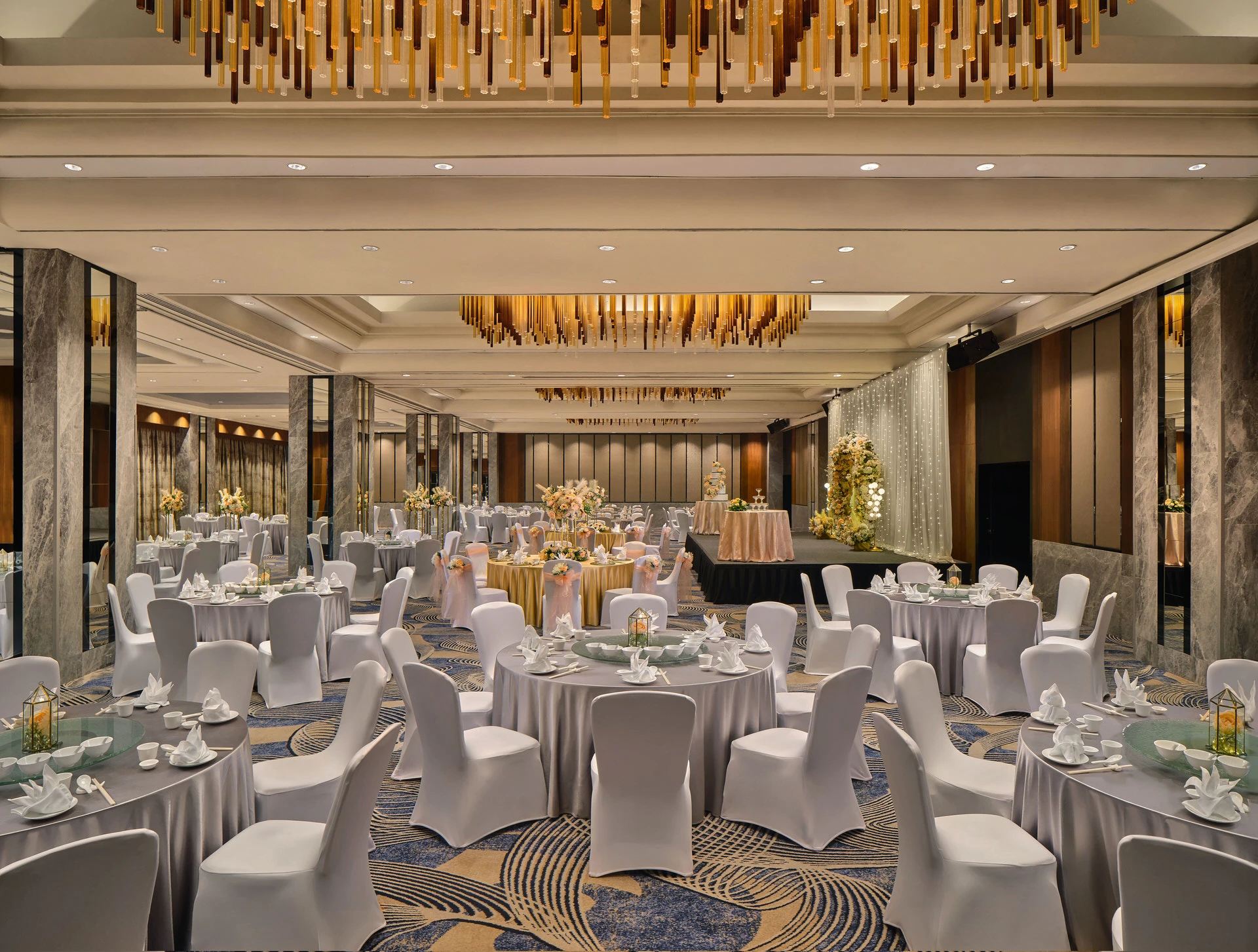 Wide view of a hotel grand ballroom with white banquet rounds, marble columns, patterned carpet, a lit stage backdrop, and gold pendant chandeliers overhead.