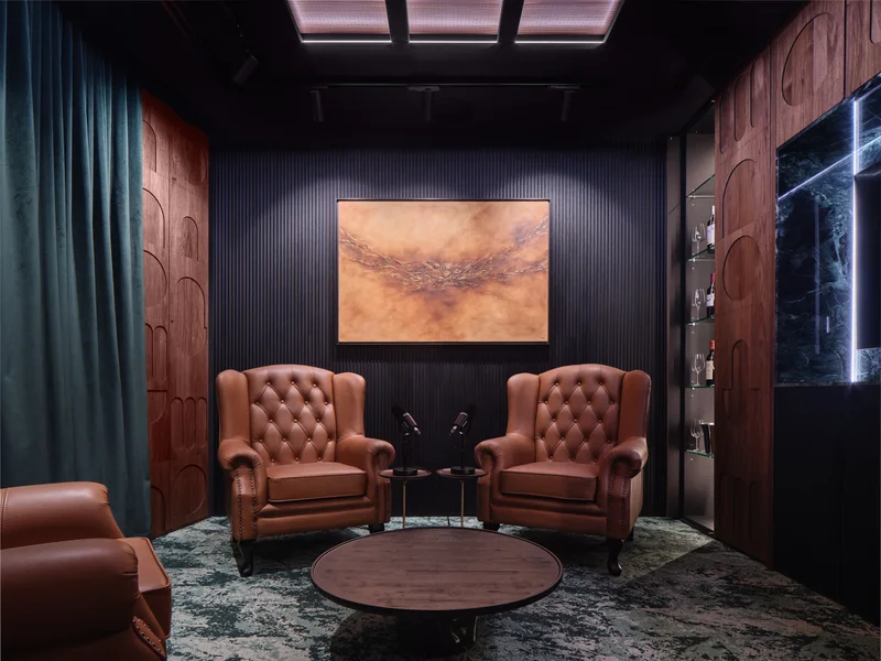 Dark executive lounge with two tufted tan leather wingback chairs, round coffee table, dark slatted accent wall, abstract painting, and teal velvet curtain.
