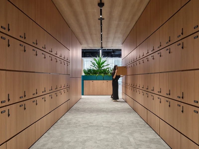 Narrow corridor lined with light timber personal lockers on both sides, a woman carrying a box at the far end, and a green planter box ahead.