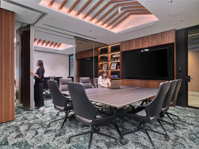 Boardroom with a long grey conference table, dark mesh chairs, large wall-mounted screen, walnut built-in shelving, slatted ceiling with pink ambient lighting.